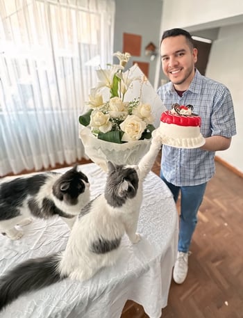 Raul with two cats, a boquet of white flowers, and a cake one of the cats is reaching for